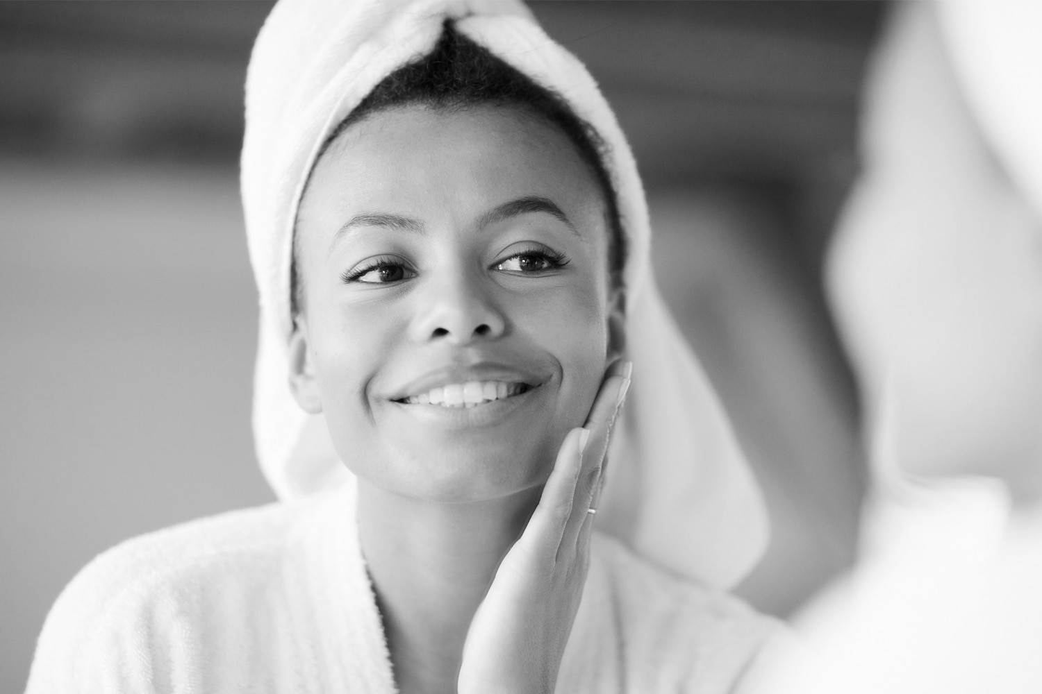 A Woman Looking In The Mirror, Smiling, With A Towel On Her Head And Her Hand On Her Face