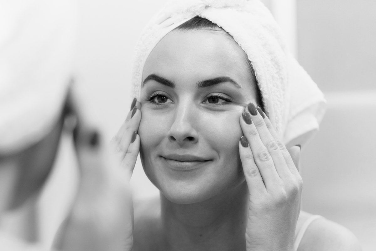 A Woman Wearing A Towel On Her Head And Smiling Into The Mirror As She Applies Something To Her Cheeks