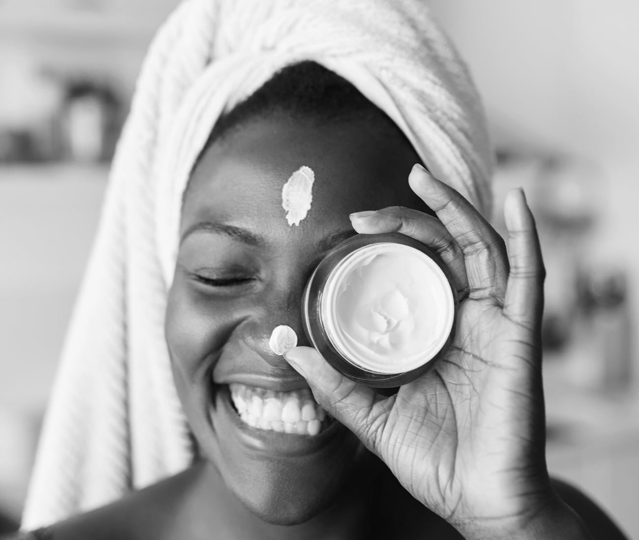 Black And White Photo Of A Woman With A Towel On Her Head, Holding Up A Cream In A Round Container, With Spots Of The Cream On Her Nose And Forehead, Smiling