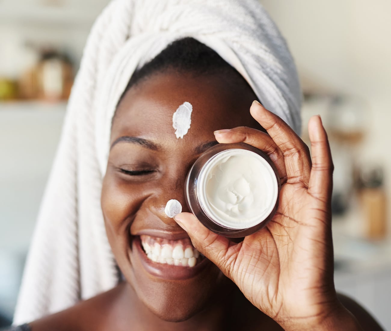 Woman With A Towel On Her Head, Holding Up A Cream In A Round Container, With Spots Of The Cream On Her Nose And Forehead, Smiling