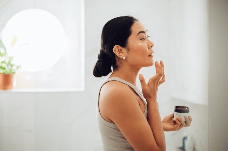 A Woman In Her Home Bathroom Applying A Skin Care Cream To Her Cheek And Studying Her Reflection