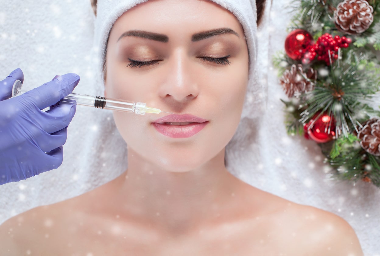 A Technician Injecting Botox Into A Female With A Santa Hat Next To Christmas Imagery.